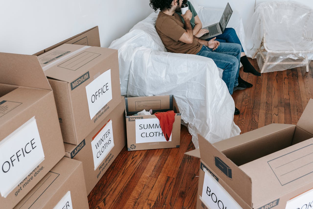 A couple seated on a couch surrounded by labeled moving boxes in a bright room, preparing to relocate.
